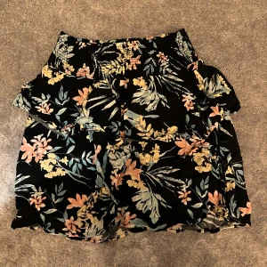 Blommig svart kortkjol med volang - Supersöt svart kortkjol med blommigt mönster i pastelliga färger. Kjolen har bred resår i midjan och volangdetaljer som ger en flowy känsla. Perfekt för sommarens alla äventyr!