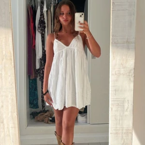 Vit kort sommarklänning med broderi - Superfin vit kort klänning med tunna justerbara axelband, v-ringning och broderade detaljer längst ner. Klänningen har en luftig passform och dragkedja i sidan. Perfekt för varma dagar och har en romantisk vibe.