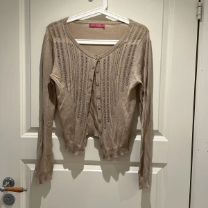 Beige kofta  - Snygg beige kofta från Indiska med lång ärm och dekorativa stickade detaljer framtill. Koftan har rund halsringning och knäppning med små knappar hela vägen. Perfekt att slänga över en topp för en chill look.