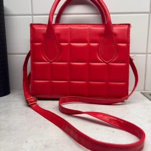 Röd quiltad axelväska med dragkedja - Snygg röd axelväska i quiltad design med rutmönster. Väskan har både handtag och justerbar axelrem samt dragkedja upptill. Perfekt storlek för det viktigaste och ger en poppig färg till din outfit.