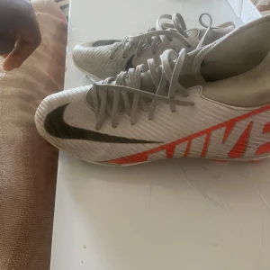 Nike fotbollsskor i vitt och röd - Snygga fotbollsskor från Nike i vit med stora svarta swoosh-loggor och röd Nike-tryck på sidan. Skorna har snörning och är tillverkade i syntetmaterial för bästa grepp och känsla på planen. Perfekta för dig som vill sticka ut med stil på matchen Ny pris 600.