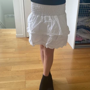 Vit volangkjol från H&M  - Supersöt vit kortkjol från H&M med bred smock i midjan och broderade detaljer. Kjolen har två lager volanger och är gjord i mjuk bomull, perfekt för en fräsch och somrig look. Storlek 140. Har en nagellacksfläck på baksidan men syns knappt.