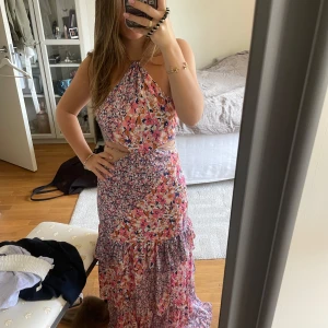 Blommig långklänning med cut-outs - Säljer en superfin långklänning med blommigt mönster i rosa, lila, vit. Klänningen har halterneck, öppna sidor med cut-outs och knytning i ryggen. Volangdetaljer nertill , perfekt för sommaren. Aldrig använd med lappen kvar!  Pris kan diskuteras!