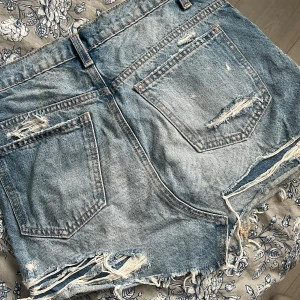 Jeanskjol från Zara med slitningar (nästan ny) - SÅ SNYGG jeanskjol från Zara, använd ett fåtal gånger. Fransiga kanter och slitna detaljer både fram och bak. Klassisk femficksdesign och cool vintagekänsla – perfekta för sommardagar och festivaler. Säljer då jag vuxit ur den.. 