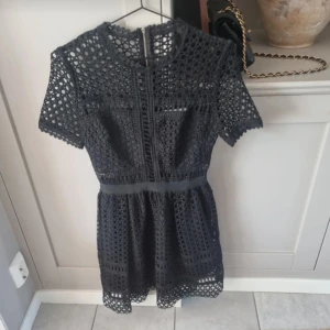 Svart kort klänning med spetsdetaljer - Supersnygg svart kortklänning med genombruten spets och rund hals. Klänningen har korta ärmar och markerad midja, perfekt för dig som gillar en trendig och feminin look. Dragkedja bak för enkel påklädning. Sparsamt använd. 