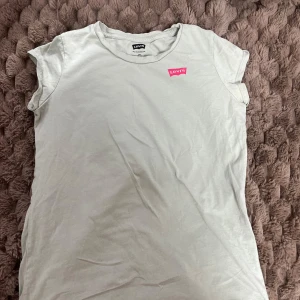 Vit t-shirt från Levi's med rosa logo - Vit t-shirt från Levi's med klassisk rund hals och korta ärmar. På bröstet sitter en rosa Levi's-logga som ger en snygg kontrast. T-shirten är i mjuk bomull och har en clean, enkel look som funkar till allt.