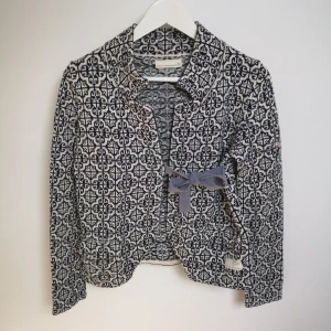 Blå/Vit stickad kofta i bomull från Odd Molly  - Blå/Vit Stickad cardigan i bommul från Odd Molly i blått och vitt med ett geometriskt mönster. Koftan har lång ärm, krage och ett knytband igråblått i midjan. Storlek 1 motsvarar Small. 