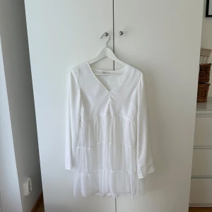 Vit kortklänning Donna Romina x NA-KD - Superfin vit kortklänning från Donna Romina x NA-KD med v-ringning, långa ärmar och volangiga lager. Klänningen har en luftig känsla och är perfekt för ljusa dagar. Materialet känns lätt och flowy, vilket ger en riktigt fräsch look.