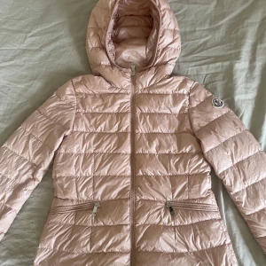 Ljusrosa dunjacka från Moncler - Supersnygg ljusrosa dunjacka från Moncler med huva och dragkedja framtill. Jackan har quiltad design, två fickor med dragkedja och klassisk Moncler-logga på ärmen. Perfekt för kyliga dagar och ger en clean look till din outfit.💘vet ej storleken tyvärr men den passade mig så skulle säga xxs! Priset går att diskutera ☺️