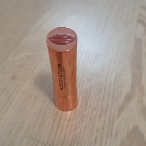 Läppstift från Revolution nude-rosa - Snyggt nude-rosa läppstift från Revolution i en lyxig roséguldig hylsa med läppmotiv på locket och själva stiftet. Krämig formula som ger en naturlig look och passar perfekt till vardags. Supersnygg detalj i sminkväskan! Har använt den men färger passar inte på mig 
