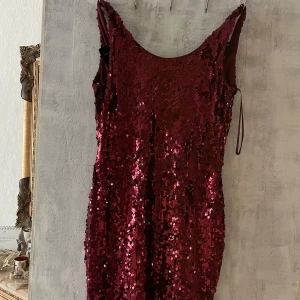 Vinröd paljettklänning med öppen rygg - Säljer en vinröd kortklänning täckt av glittriga paljetter. Klänningen är ärmlös och har en snygg öppen rygg med dekorativa svarta pärlor. Perfekt för dig som vill sticka ut och glänsa på festen!