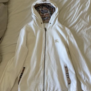 Vit hoodie från Burberry - Vit hoodie från Burberry med klassiskt rutigt foder i huvan och på fickorna. Jackan har dragkedja framtill, lång ärm och ikonisk Burberry-logga broderad på bröstet.