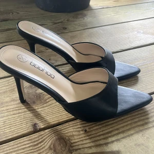 Svarta klackpumps från Boohoo - Stilrena svarta pumps från Boohoo med spetsig tå och hög klack. Skorna har öppen häl och bred rem över foten, vilket ger en modern och clean look. Tillverkade i syntetmaterial med en elegant finish. Perfekta för dig som vill sticka ut med en klassisk men trendig vibe.