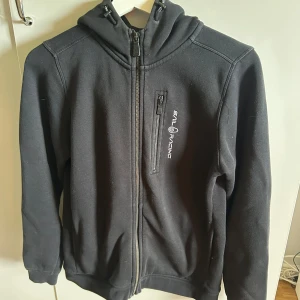 Svart hoodiejacka Sail Racing 160 - Svart hoodiejacka från Sail Racing med dragkedja framtill och bröstficka med logga. Mjuk insida och huva med snörning. Tillverkad i bomull och polyester, perfekt för chill dagar eller när du vill ha en sportig look. Den är stor i storleken så den passar även som en XS!