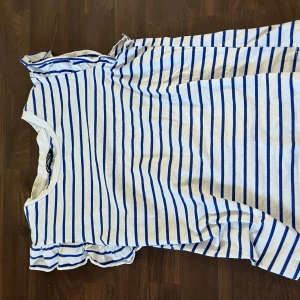 Blus - Säljer en vit och blå ärmlös t-shirt med smala ränder och volangdetalj vid axlarna. T-shirten har en klassisk rund halsringning och är gjord i mjuk bomull, perfekt för en chill och fräsch look.den är från SHEIN 