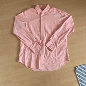 Rosa skjorta från Ralph Lauren  - Snygg rosa skjorta från Ralph Lauren i storlek L. Klassisk modell med krage, knappar hela vägen och broderad logga på bröstet. Skjortan är långärmad och tillverkad i bomull, perfekt för en clean och stilren look. Väldigt trendig och generellt väldigt snygg.