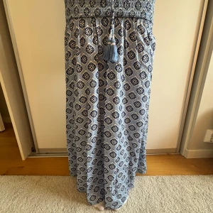 Söt lå långklänning med mönster - Luftig långklänning i ljusblått och mörkblått med grafiskt mönster. Smockad överdel och dekorativ snörning med tofsar i midjan. Perfekt för varma dagar och har en avslappnad, bohemisk vibe.