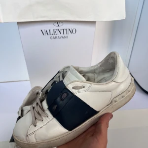 Valentino opens - Snygga vita sneakers från Valentino Garavani med bred mörkblå läderdetalj över snörningen. Klassisk rund tå, platt sula och grå snören. Skorna är tillverkade i skinn och har en stilren, modern look som passar till det mesta. Enda negativa är hälen i insidan