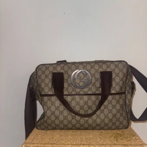 Gucci brun axelväska med GG-mönster - Säljer en snygg axelväska från Gucci i beige och brunt med klassiskt GG-mönster över hela väskan. Väskan har stora dubbla G-loggan framtill, bruna handtag och justerbar axelrem. Tillverkad i canvas med detaljer i skinn och dragkedja upptill.
