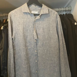 Grå  linneskjorta  - Säljer en stilren grå skjorta från S.C.W. i 100% linne. Skjortan har klassisk krage, knäppning framtill och långa ärmar. Skjortan är helt ny med lapp på!!