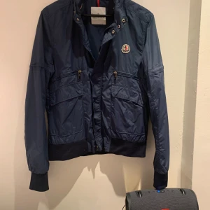 Blå vindjacka från Moncler - Säljer en snygg blå vindjacka från Moncler med dragkedja och ribbade muddar. Jackan har  flera praktiska fickor med dragkedja. Perfekt för en stilren look. luva kommer med jackan som man kan stoppa in och ta ut 