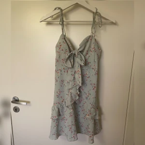 Blommig klänning - Söt blommig klänning i turkos nyans med volangdetaljer och knytband framtill. Endast använd två gånger. Klänningen har smala axelband som man knyter. Den har rosa blommönster och är perfekt för sommaren. Den har en dragkedja baktill och är från Cider i storlek xxs. Har ni några frågor är det bara att skriva! 💕