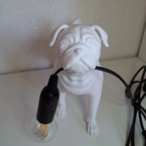 Vit hundformad bordslampa - Unik bordslampa i form av en vit hund som håller en glödlampa i munnen. Lampan har en modern och stilren design, perfekt för att ge en lekfull touch till ditt rum. Tillverkad i plast. med en svart sladd.  Mått ca: 20,5 cm hög 