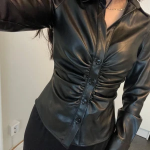 Svart skinnskjorta med knappar - Snygg svart skinnskjorta med knappar och rynkdetaljer framtill. Skjortan har långa ärmar och en figurnära passform som ger en stilren look.