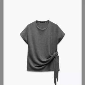 Grå topp med knyt från Zara - Snygg grå topp från Zara med korta ärmar och en stilren knytning på sidan. Perfekt för en chic och avslappnad look. Tillverkad i ett mjukt material som ger en bekväm passform.