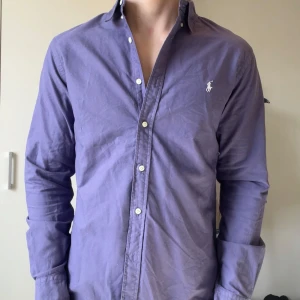 Lila skjorta från Ralph Lauren - Snygg lila skjorta från Ralph Lauren med klassisk krage och broderad logga på bröstet. Skjortan har långa ärmar och knappar framtill. Perfekt för en stilren look!