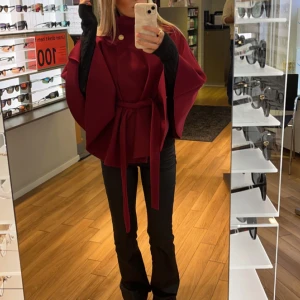 Vinröd kappa/poncho med knytband - Elegant vinröd kappa med knytband i midjan och dekorativa knappar vid halsen. Den har en lös passform och är perfekt för höst och vår. Kappan har en stilren design som ger en sofistikerad look. Storlek: onesize