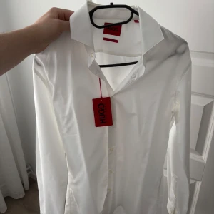 Vit skjorta från Hugo Boss - Elegant vit skjorta från Hugo Boss med klassisk krage och knappar framtill. Perfekt för en stilren look. Skjortan har långa ärmar och är tillverkad i ett mjukt material för extra komfort. Helt ny 