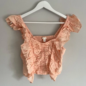 Persikofärgad blus från H&M - Säljer en söt persikofärgad topp från H&M med volanger och broderade detaljer. Blusen har en smockad design och korta ärmar med volang. Perfekt för en somrig look! 🌸