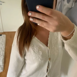 Vit kofta med broderade blommor - Säljer en söt vit kofta med broderade blommor framtill. Koftan har långa ärmar och knappar i trälook. Perfekt för en stilren och romantisk look.