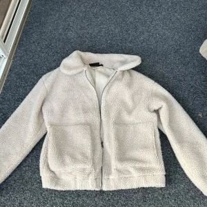 Beige teddyjacka från Tindra - Mysig beige teddyjacka från Tindra med dragkedja framtill. Jackan har en fluffig textur och långa ärmar, perfekt för kyligare dagar. Den har en enkel design med två fickor framtill och en bekväm passform.