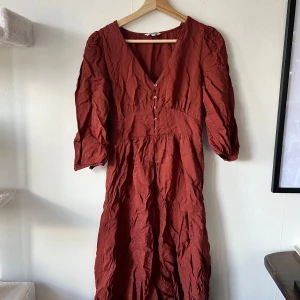 Rostfärgad klänning från Indiska - Säljer en vacker rostfärgad klänning från Indiska med v-ringning och knappar framtill. Klänningen har trekvartsärmar och en avslappnad passform. Perfekt för höst och vår!