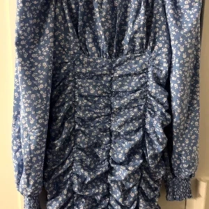 Blå blommig klänning - Charmig blå klänning med små vita blommor och rynkade detaljer. Klänningen har långa ärmar med elastiska muddar och en smickrande passform. Perfekt för en somrig look! Strl. L