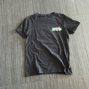 Svart t-shirt från Billabong - Säljer en svart t-shirt från Billabong med färgglatt tryck på både fram- och baksidan. T-shirten är kortärmad och tillverkad i 100% bomull. Perfekt för en avslappnad stil med en touch av färg. Pris kan diskuteras. 