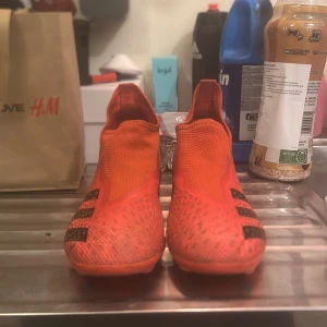 Adidas Predator fotbollsskor - Snygga och sportiga Adidas Predator fotbollsskor i en iögonfallande orange färg med svarta detaljer. Skorna har en strumpliknande passform utan snörning och är designade för att ge bra grepp på planen. Perfekta för fotbollsspelare som vill ha både stil och funktion.