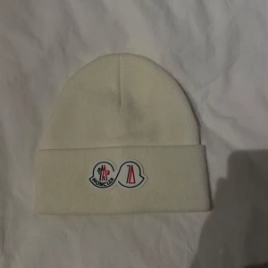 Vit mössa från Moncler - Snygg vit mössa från Moncler med deras ikoniska logotyp framtill. Tillverkad i 100% akryl för en mjuk och bekväm känsla. Perfekt för att hålla värmen under kyliga dagar.