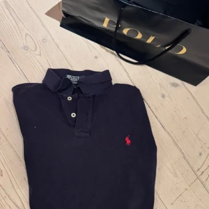 Svart kortärmad pikétröja från Polo - Säljer en klassisk svart pikétröja från Polo med korta ärmar och en röd broderad logga på bröstet. Perfekt för en stilren och avslappnad look. Tröjan har knappar vid halsen för en justerbar passform.