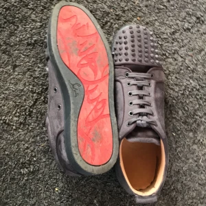 Grå sneakers med nitar från Christian Louboutin - Snygga grå sneakers från Christian Louboutin med ikonisk röd sula och nitar på ovansidan. Skorna har snörning och är tillverkade i mocka för en lyxig känsla. Perfekta för att ge din outfit en edgy touch.