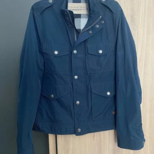 Mörkblå field- jacka från Burberry - Säljer en stilren mörkblå jacka från Burberry med flera fickor och knappar. Jackan har en klassisk design med axelklaffar och dragkedja framtill. Perfekt för en elegant look. Nypris 9000 kronor. Öppen för prisförslag. Aldrig använd. Passar 182-190. 