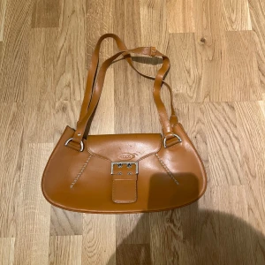 Tod’s Handväska - Säljer en Brun/orange handväska med silverfärgade detaljer som gör den väldigt elegant. Väskan är från det italienska märket Tod’s. Väldigt rymlig och enkel att bära. Perfekt till sommaren. Hör av er vid intresse eller frågor. PRIS KAN DISKUTERAS!