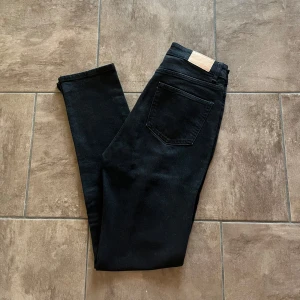Svarta jeans med slits  - Snygga svarta jeansbyxor med hög midja och raka ben. Byxorna har klassisk femficksdesign och silverfärgade knappar och även en slits på benet nedtill. Perfekta för en stilren look. Storlek 38, normala i storlek. Midjemått: 36cm innerbenslängd: 79cm