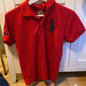 Röd pikétröja med broderad logga - Snygg röd pikétröja från Ralph Lauren med en stor broderad logga i marinblått på bröstet och siffran 3 på ärmen. Perfekt för en stilren och sportig look.