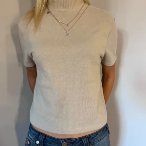Beige ribbad polotröja - Säljer en stilren beige ribbad polotröja med korta ärmar. Perfekt för en avslappnad men ändå elegant look. Tröjan har en hög krage och är tillverkad i ett mjukt material.