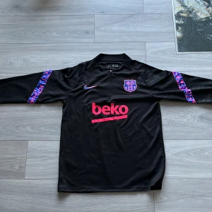 Svart fotbollströja från Nike - Säljer en svart fotbollströja från Nike med FCB-logga och sponsortryck i rosa. Tröjan har långa ärmar med ett färgglatt mönster på ärmarna och en halv dragkedja framtill. Perfekt för fotbollsfans! ⚽