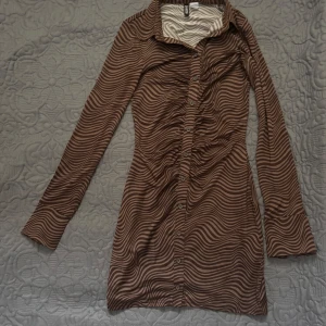 Brun randig klänning från H&M Divided - Snygg brun klänning med vågiga ränder från H&M Divided. Klänningen har långa ärmar och knappar framtill. Tillverkad i mjukt polyester med lite stretch för en bekväm passform. Priset kan diskuteras!