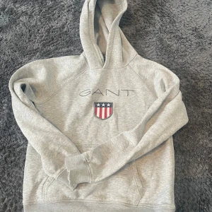 Grå hoodie från GANT - Säljer en stilren grå hoodie från GANT  som inte säljs längre där jag köpte för 500 med deras ikoniska logga och ett sköldmärke på bröstet. Tröjan har en klassisk känguruficka och en bekväm huva. Perfekt för en avslappnad stil.